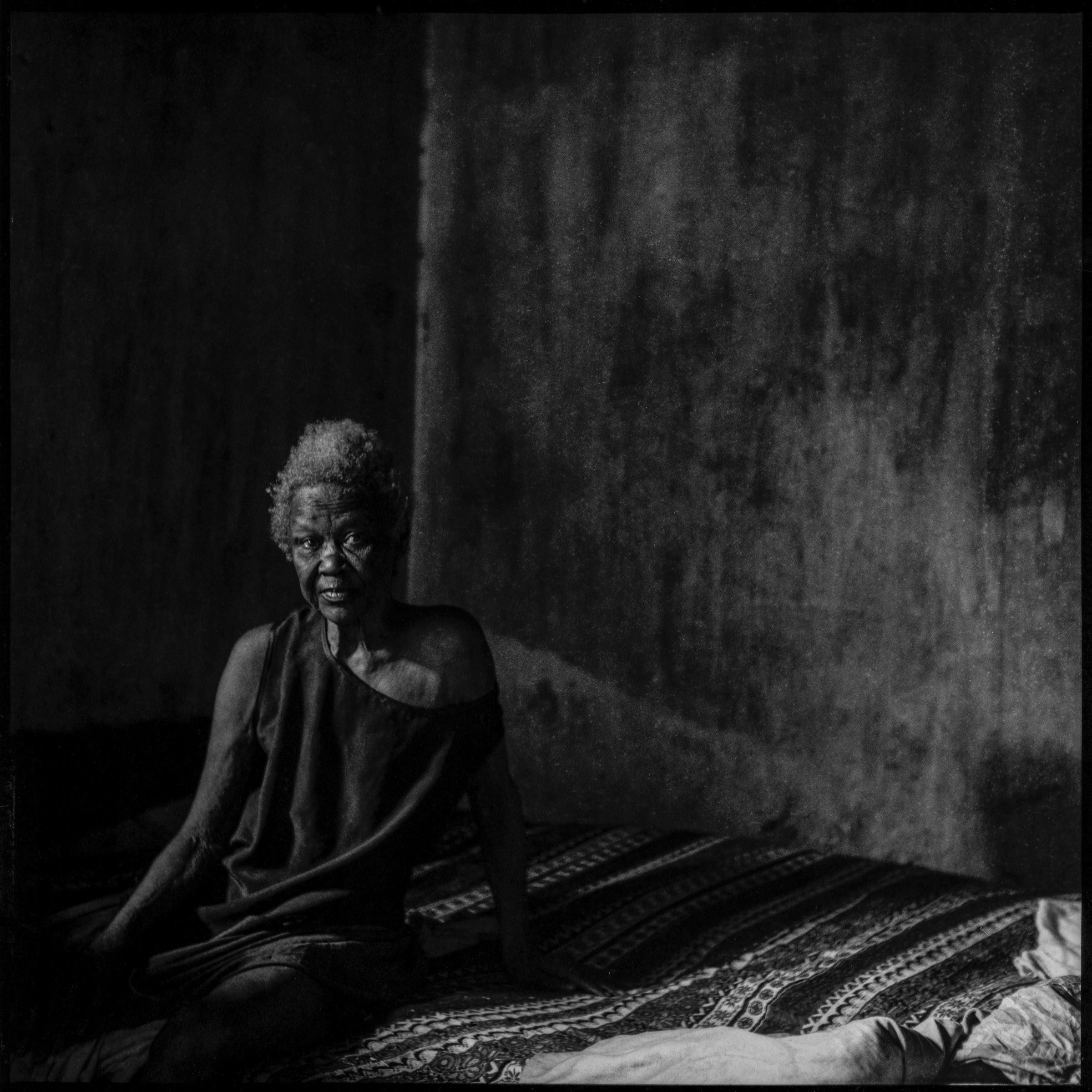 A seated woman beside a textured wall, photographed in black and white.
