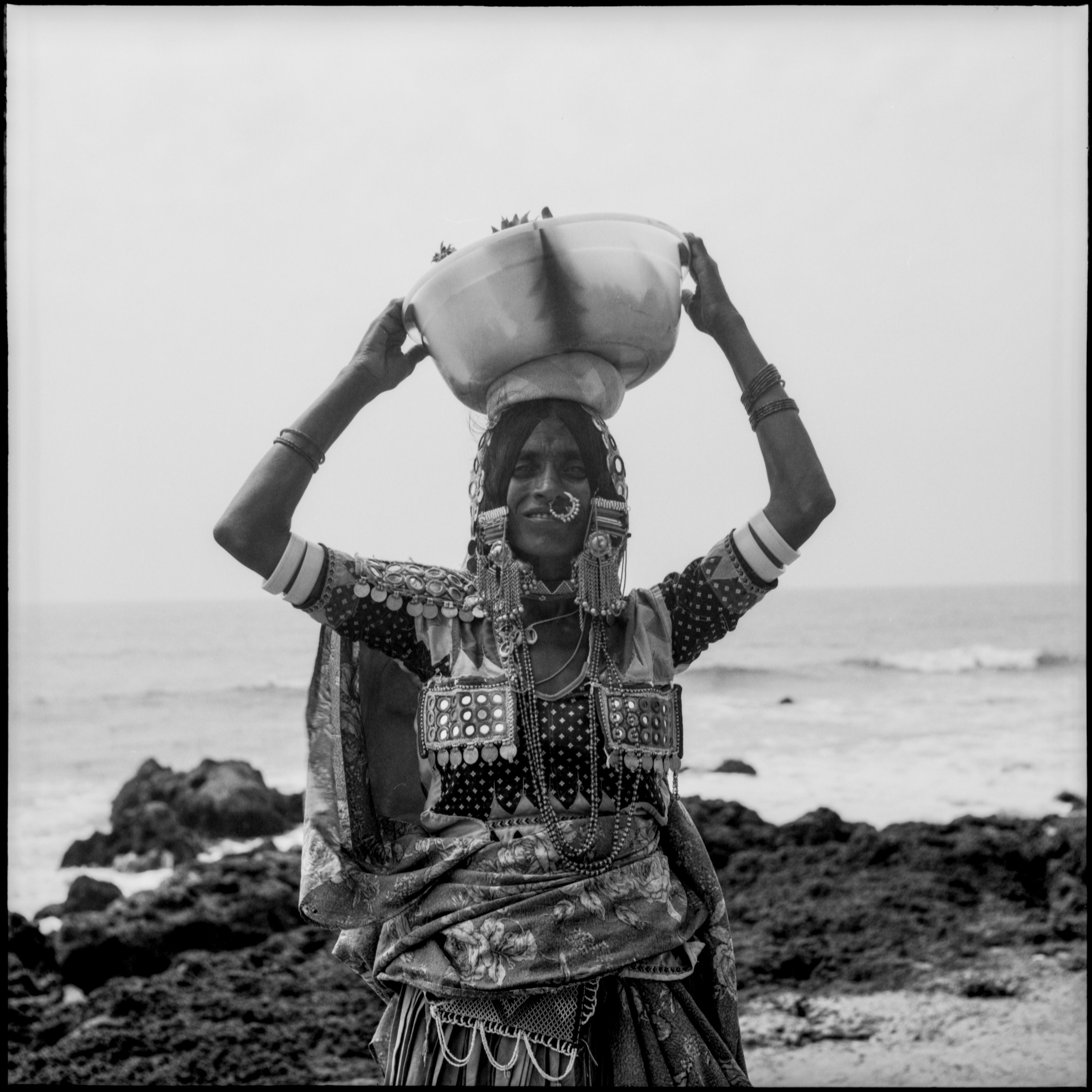 A woman carrying a bowl on her head beside the sea, photographed in black and white.