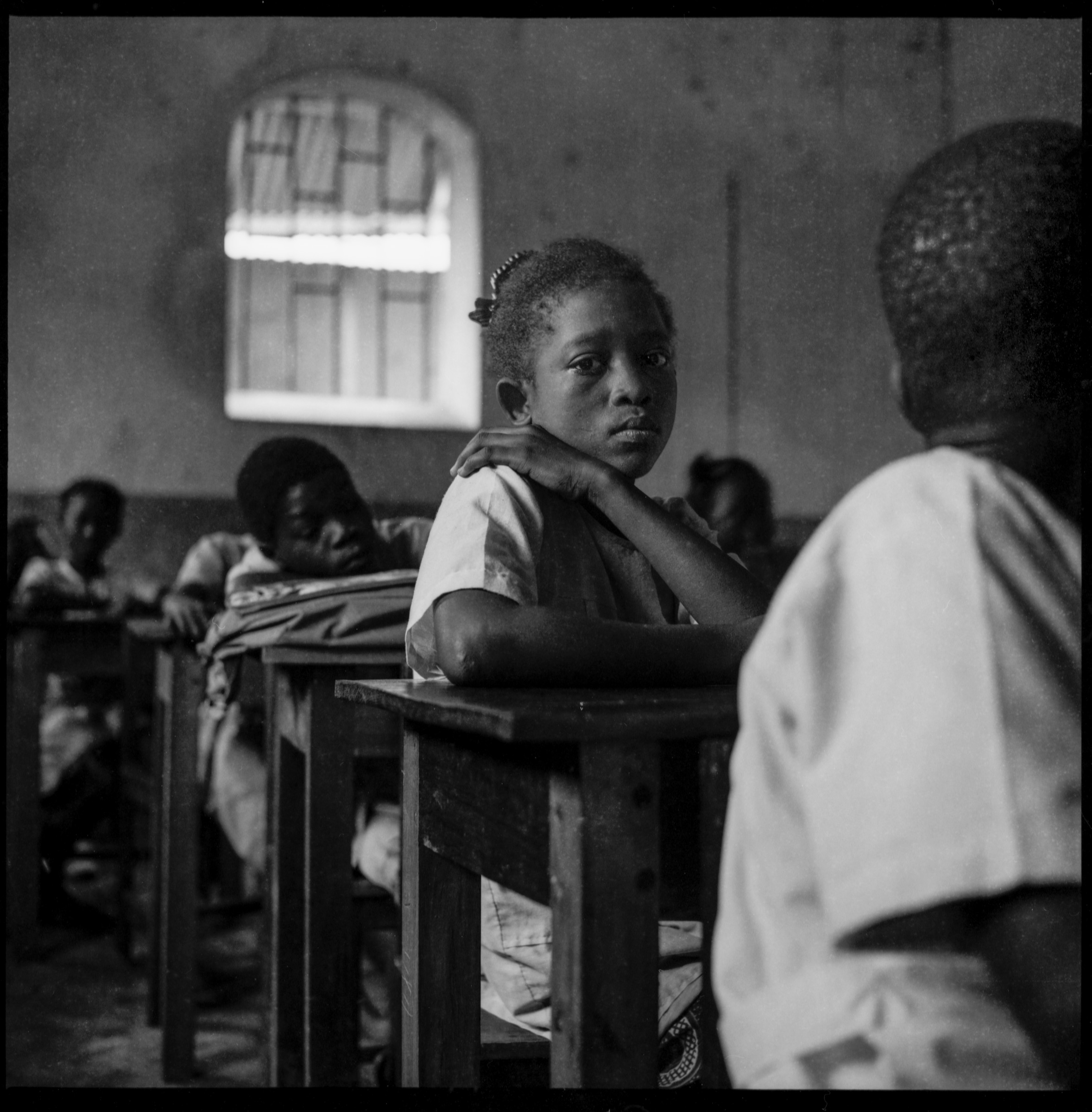A pupil looking toward the camera across a classroom, photographed in black and white.
