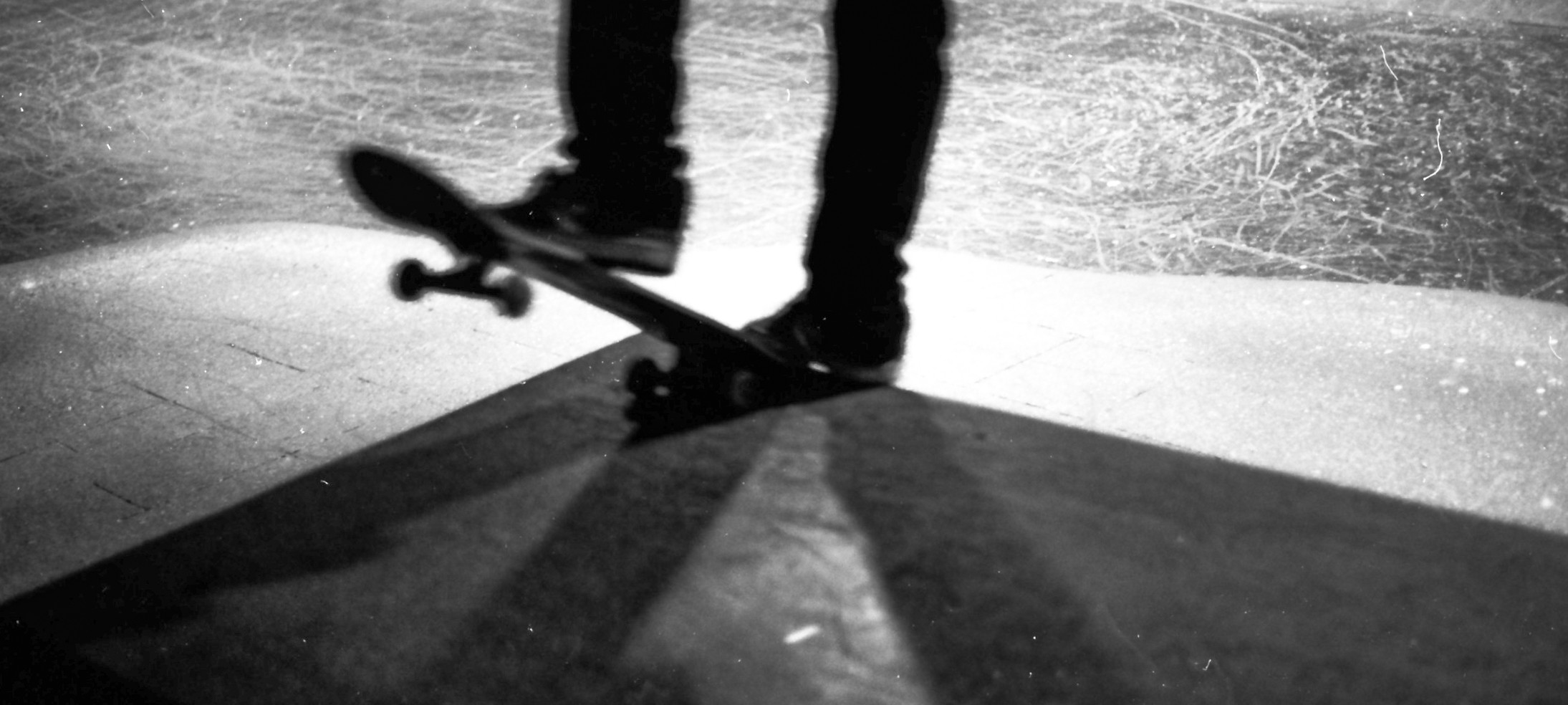 A skateboard and feet crossing geometric shadows on a ramp in black and white.