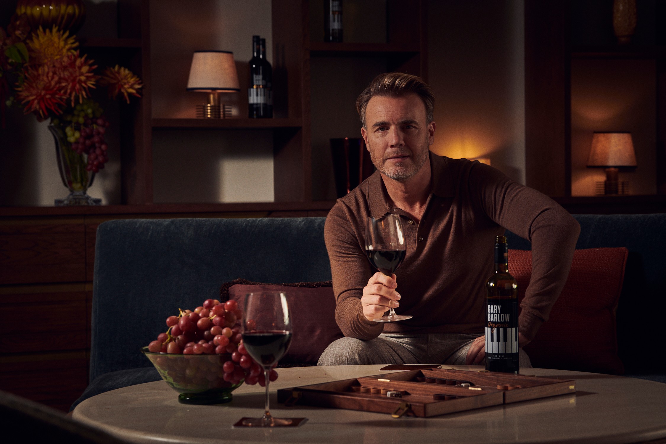 Gary Barlow seated with a glass of wine in a warmly lit interior.